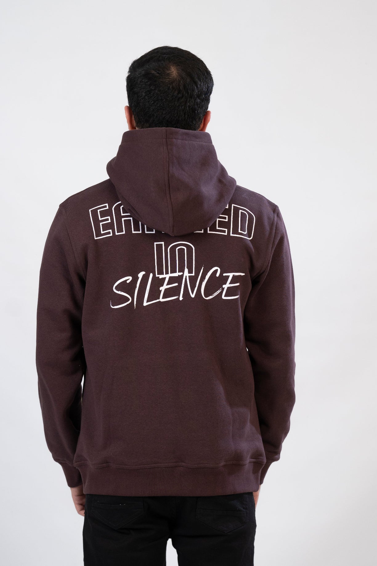 Person wearing a dark hoodie with 'EARNED IN SILENCE' text on the back against a white background