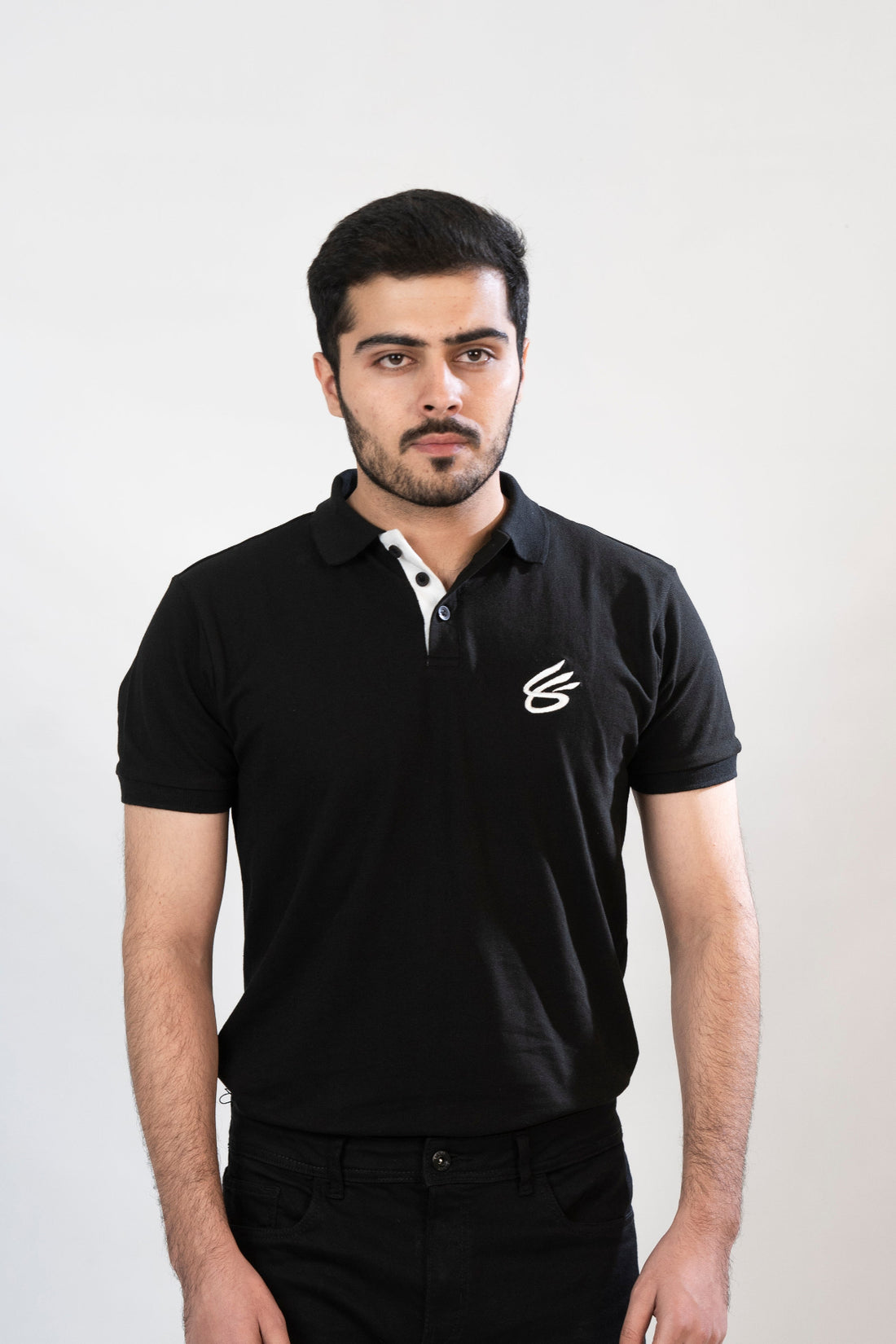 A person wearing a black polo shirt with a white collar and a logo on the chest, standing against a neutral background.