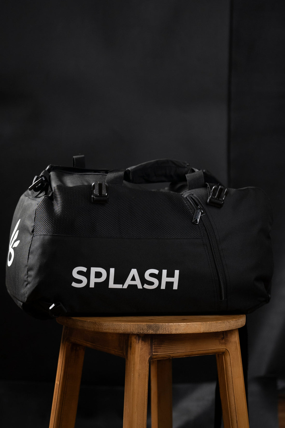 A black Splash duffle bag with the brand name printed on the side, placed on a wooden stool against a dark background.