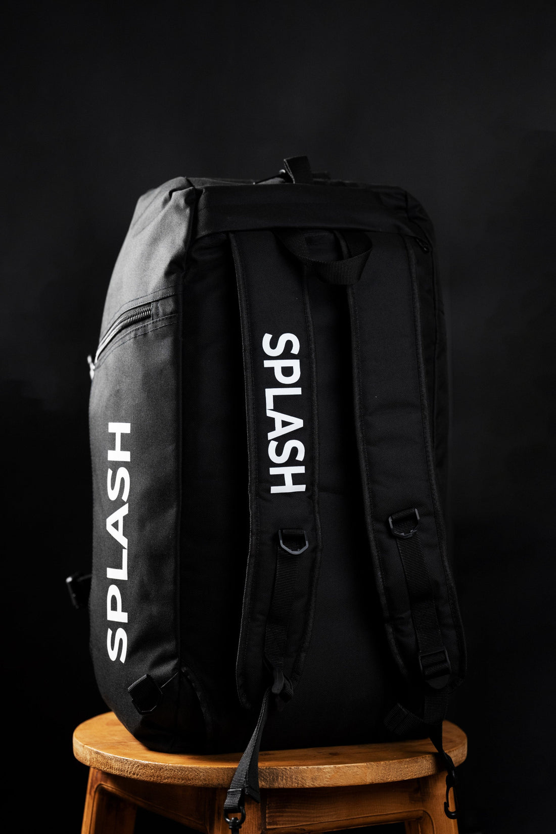 A black Splash duffle bag with the brand name printed on the side, placed on a wooden stool against a dark background.
